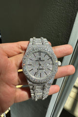 A heavily paved silver-tone watch with a matching paved bracelet, held in a person's hand. The watch features a silver-tone case, a date window at 3 o'clock, and a date-day bezel with a diamond-encrusted dial.